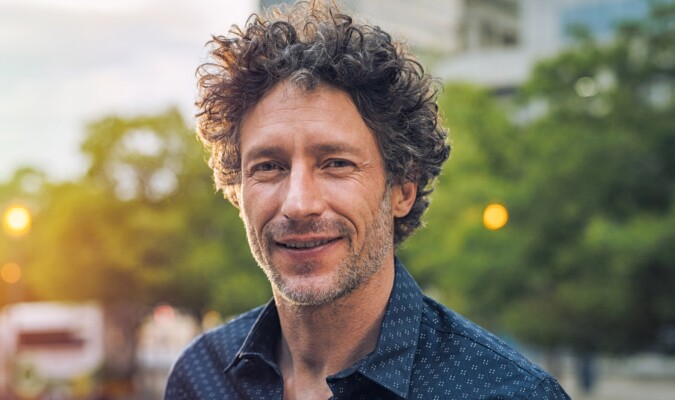 A middle-aged man with brown curly hair in a navy polo smiling at the camera with trees in background.